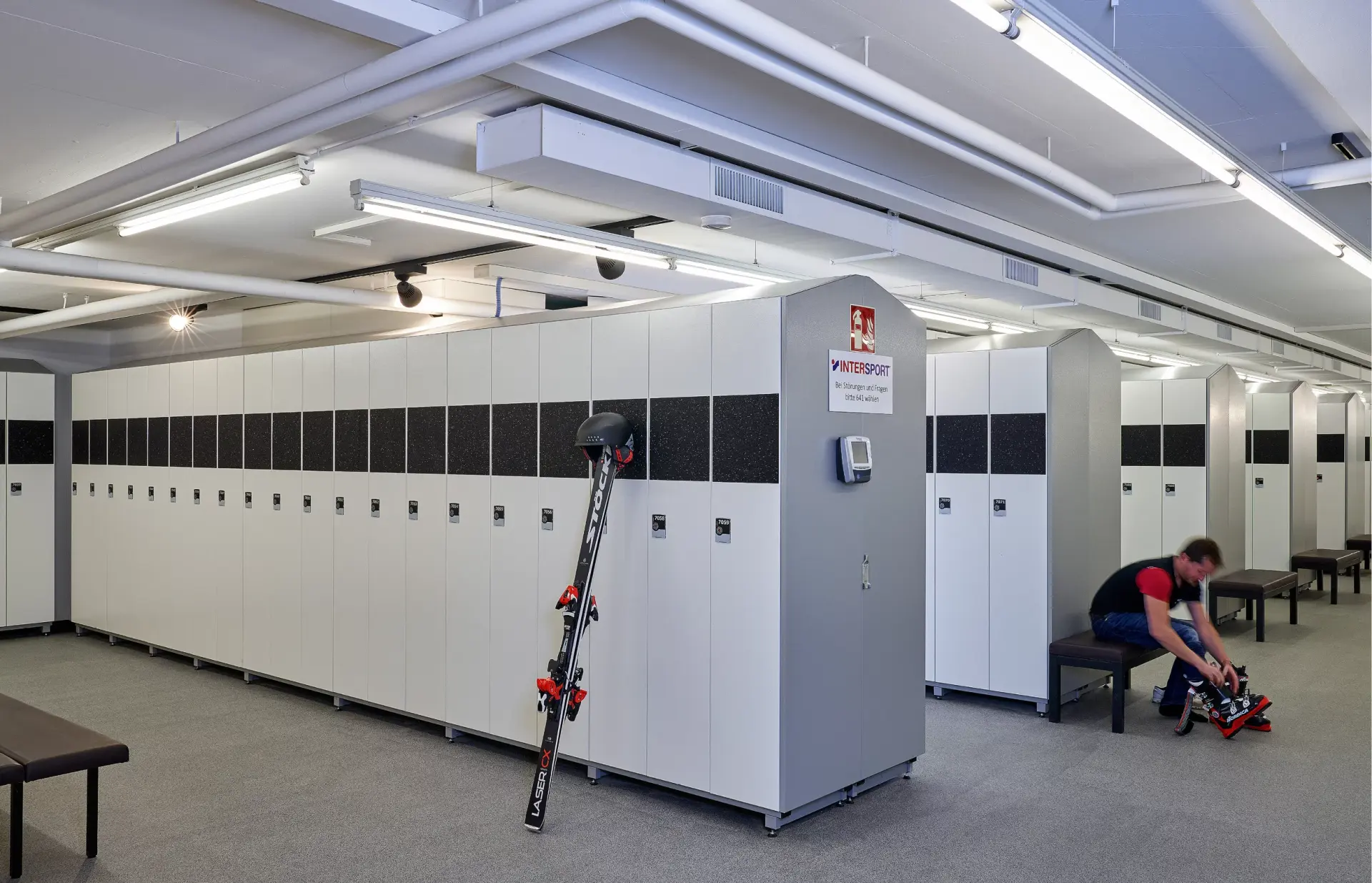 A man sits on a bench in a ski locker room, wearing black pants and a red and black shirt, adjusting ski equipment. White lockers line the walls under bright lighting.