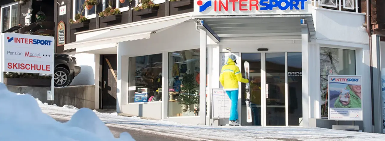 Winter scene of an Intersport shop with a ski school sign. A person in bright ski attire stands outside. Snow decorates the path and roof.