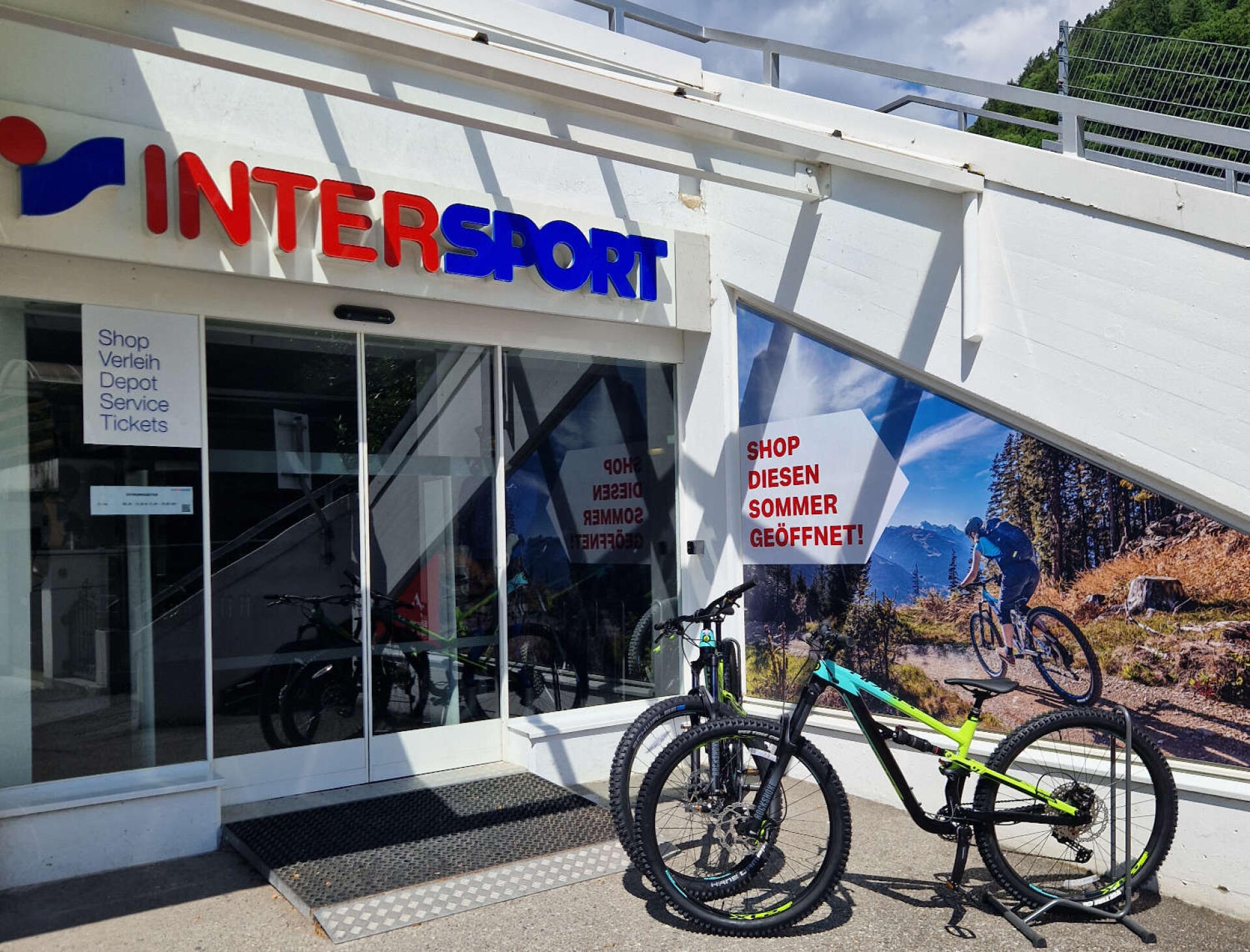 Intersport-Ladenfront mit Fahrrädern, die draußen ausgestellt sind. Ein Mountainbike-Poster ist zu sehen. Die Umgebung ist sonnig und vermittelt ein lebendiges und aktives Gefühl.