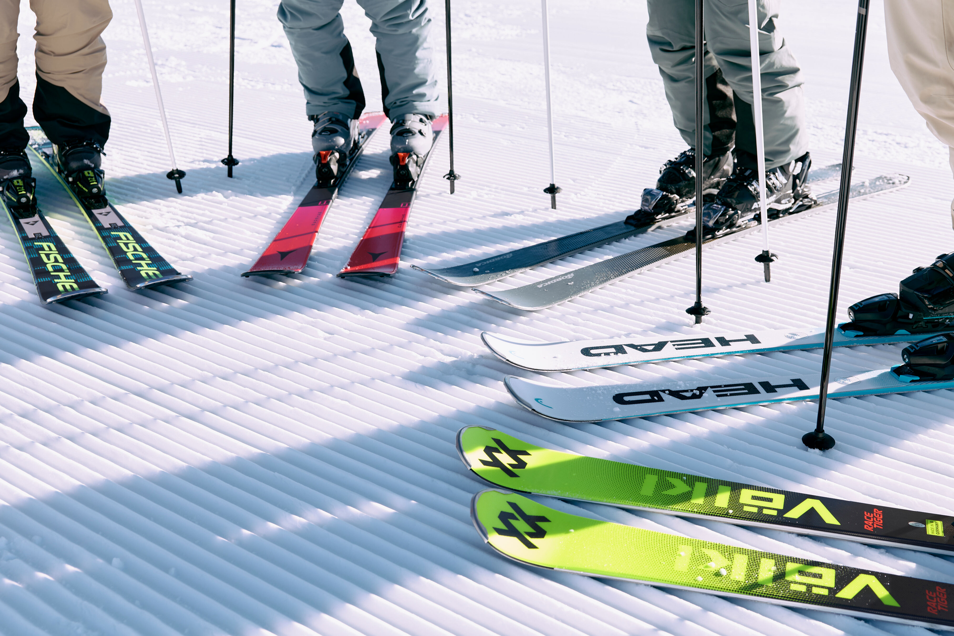 A skier in an orange jacket holds skis on a snowy mountain. Four colorful ski pairs are displayed on the left. Blue sky and peaks in the background. Energetic mood.