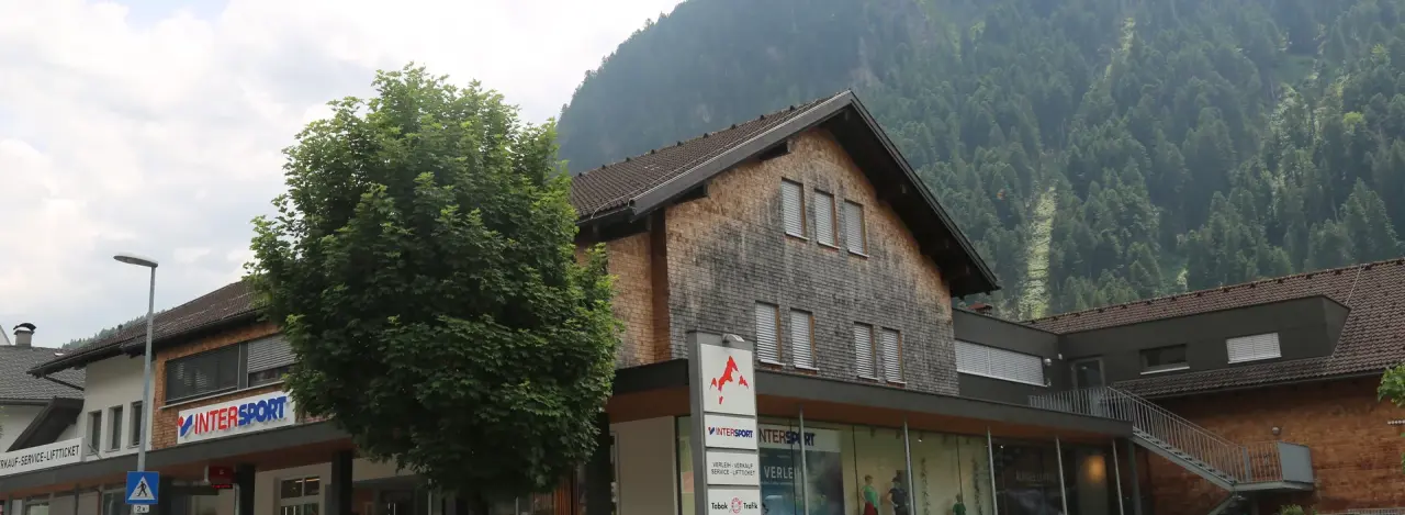 Ein schneebedecktes Gebäude im Chalet-Stil mit Intersport-Beschriftung vor dem Hintergrund schneebedeckter Berge und einem klaren blauen Himmel, das eine winterliche, ruhige Atmosphäre vermittelt.