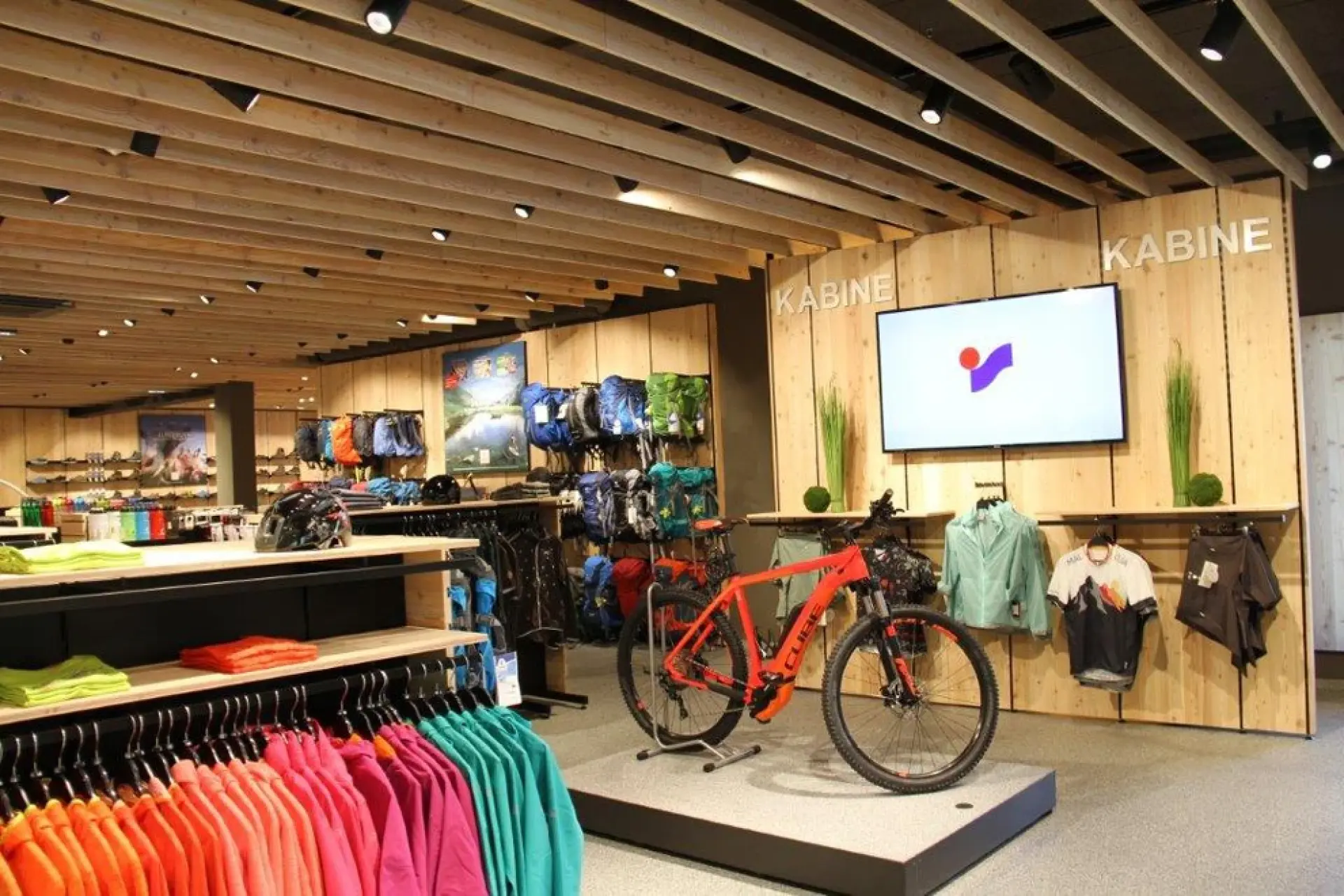 Modernes Interieur eines Intersport-Geschäfts mit Holzdecken und farbenfroher Kleidung auf Ausstellungsständern. Ein rotes Fahrrad steht unter warmer Beleuchtung prominent im Vordergrund.