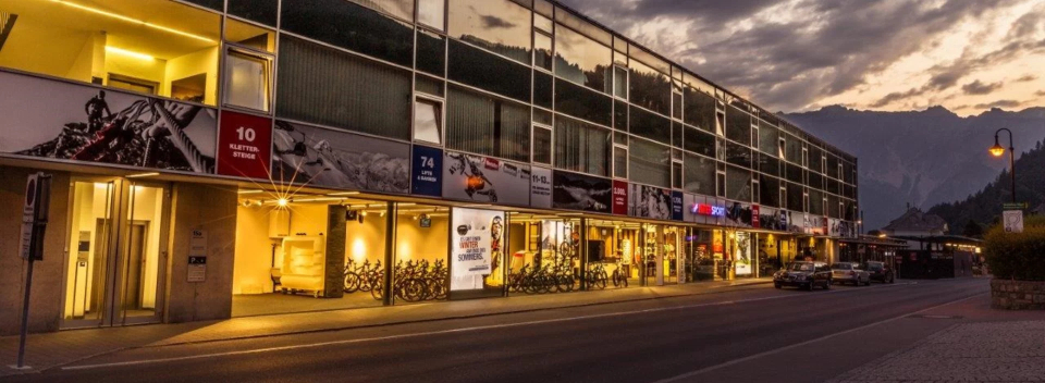 Abendliche Straßenszene mit einem beleuchteten modernen Gebäude mit Ladenfronten und bunten Schildern. Berge und ein bewölkter Himmel bilden den Hintergrund und schaffen eine gemütliche Atmosphäre.