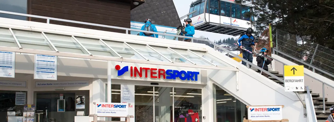 Außenaufnahme mit einem Intersport-Geschäft unterhalb einer Treppe und einer Seilbahn oberhalb. Skifahrer in blauen Outfits gehen die Treppe hinunter.