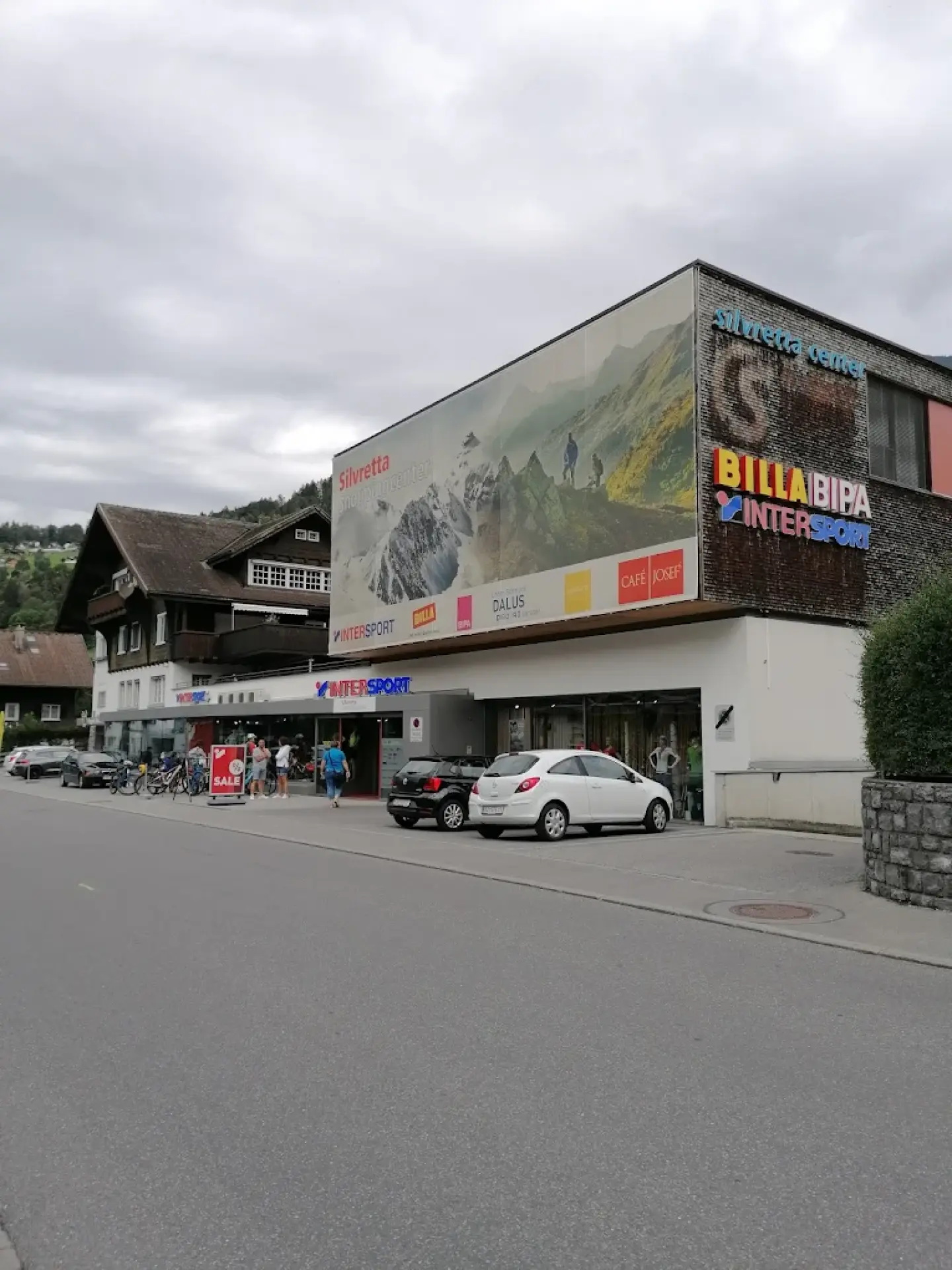 Straßenszene mit einem Gebäude, das mit einem großen Wandgemälde und Werbeplakaten verziert ist. In der Nähe stehen Autos geparkt. Der Himmel ist bewölkt, was eine bedrückte Atmosphäre schafft.