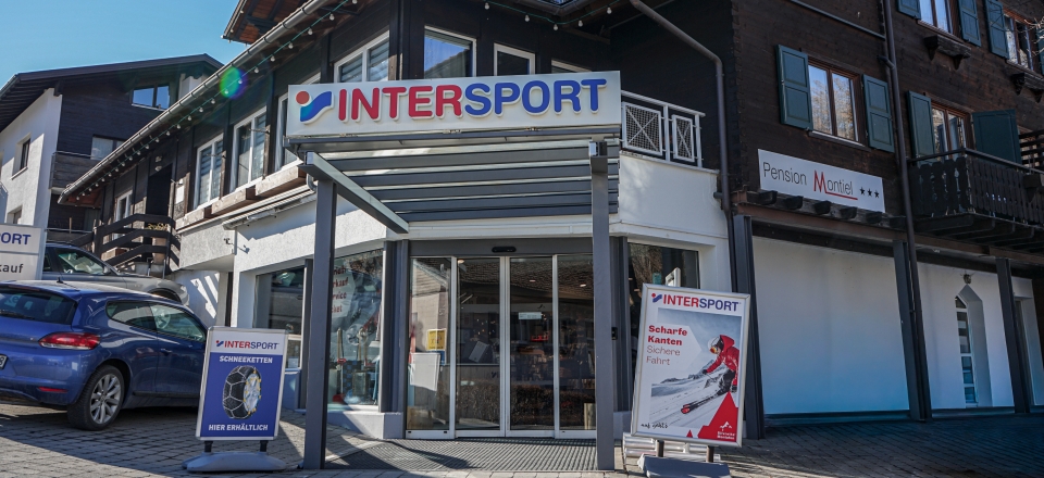 Winterszene vor einem Intersport-Geschäft mit einem Schild einer Skischule. Eine Person in heller Skikleidung steht draußen. Schnee bedeckt den Weg und das Dach.