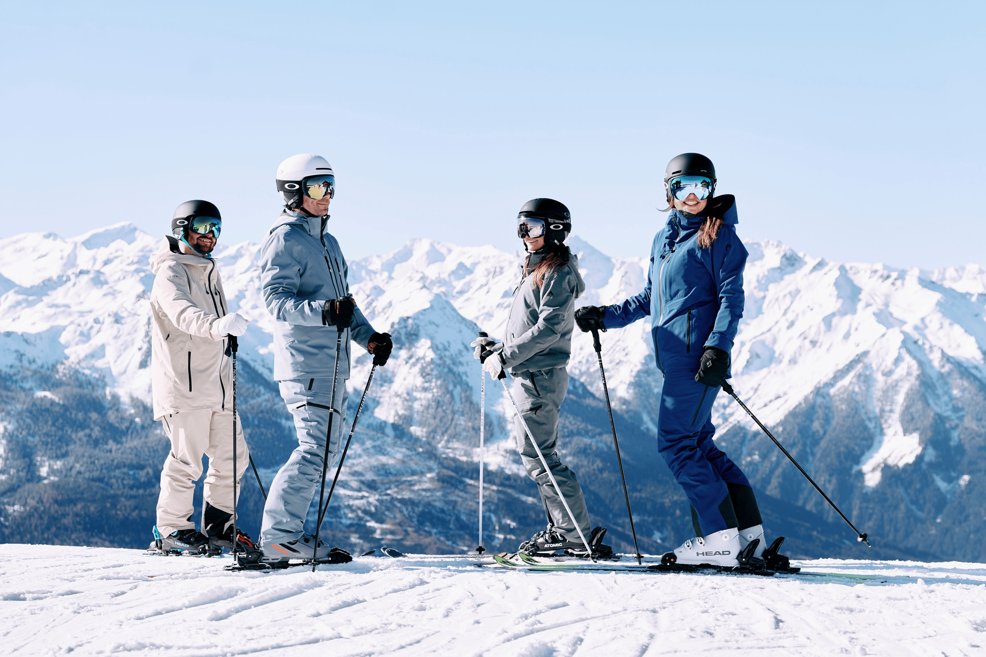 Zwei Skifahrer in leuchtender Kleidung fahren einen verschneiten Berghang hinunter, umgeben von weitläufigen weißen Landschaften und fernen Bergen unter einem klaren blauen Himmel.
