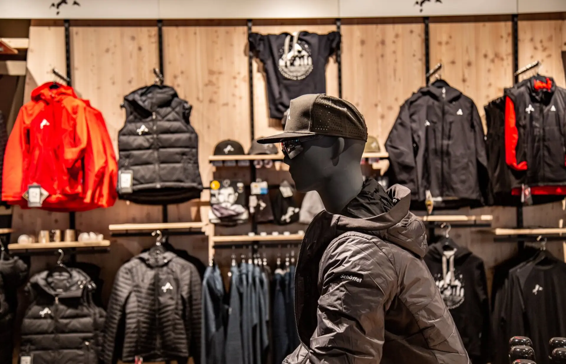 Mannequin mit grauer Jacke und Mütze in einem Bekleidungsgeschäft, umgeben von verschiedenen Jacken und Hemden in Rot- und Schwarztönen. Warme, gemütliche Atmosphäre.