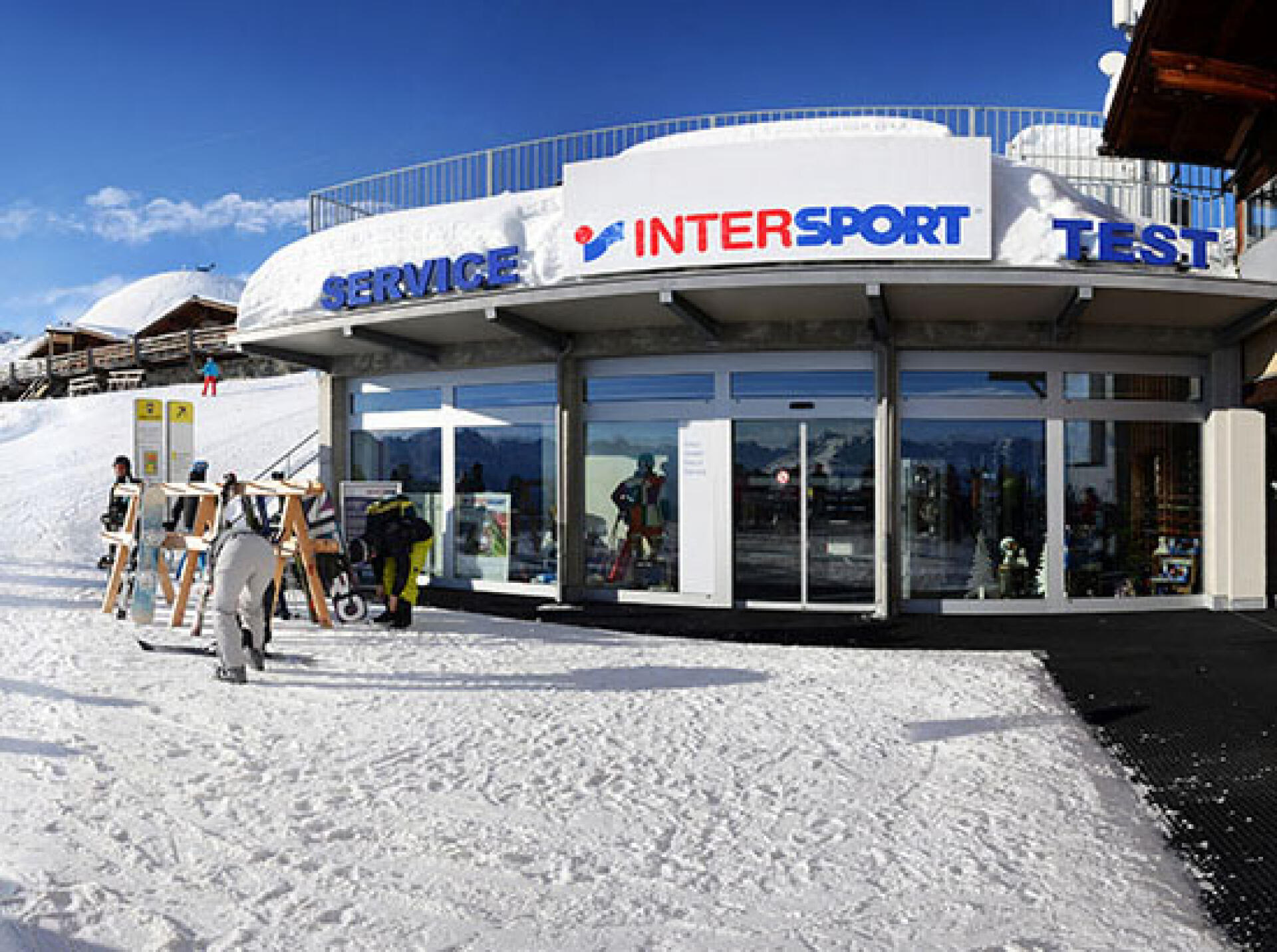 Ein schneebedeckter Intersport-Laden mit Glasfassade steht in einem Skigebiet unter strahlend blauem Himmel. Skier und Stöcke sind draußen ausgestellt, was eine lebendige, aktive Atmosphäre suggeriert.