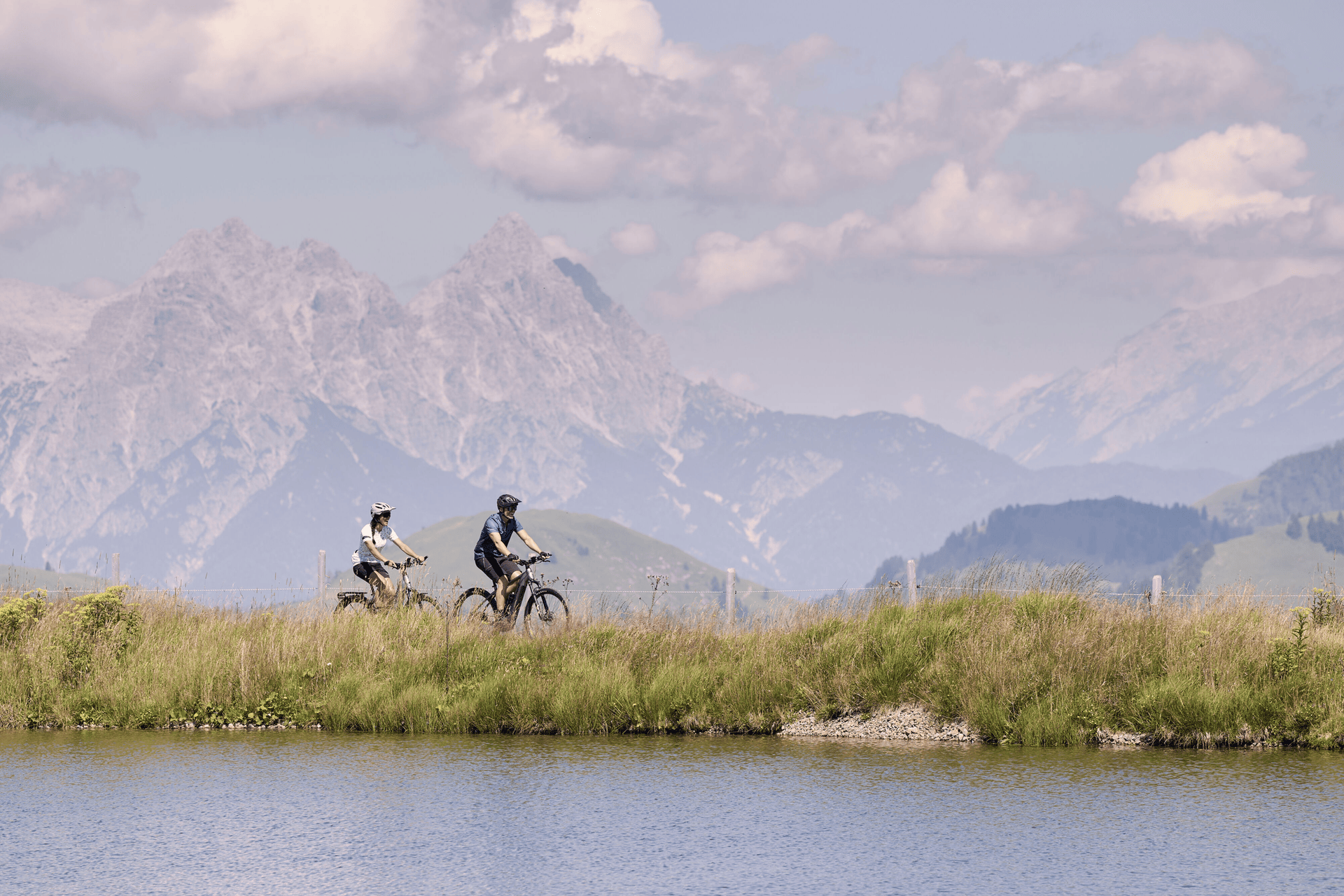 Drei Radfahrer fahren mit Mountainbikes auf einem grasbewachsenen Hügel unter einem strahlend blauen Himmel mit flauschigen Wolken. Im Hintergrund erheben sich majestätische Berge.