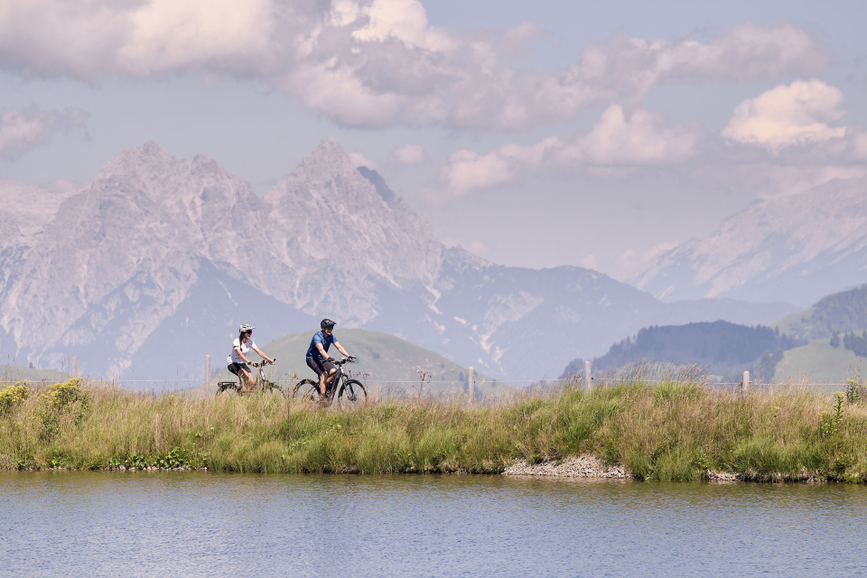 Three cyclists ride mountain bikes on a grassy hilltop under a bright blue sky with fluffy clouds. Majestic mountains rise in the background.