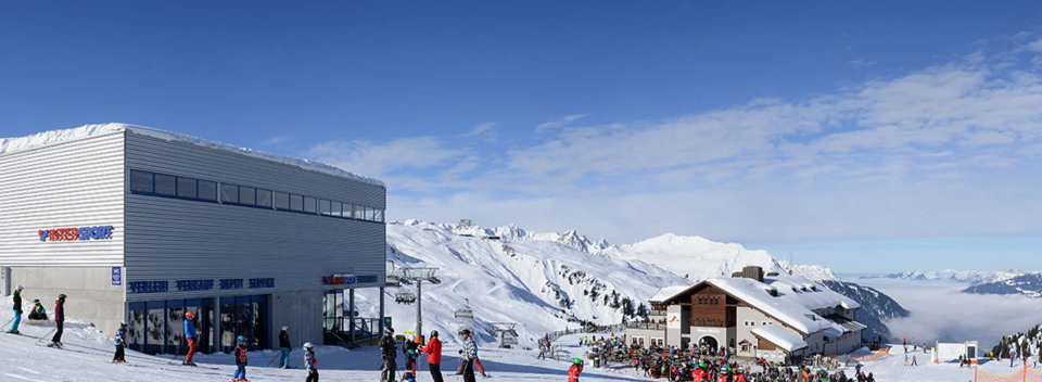 Ein verschneites Skigebiet in den Bergen mit regem Treiben. Menschen in farbenfroher Winterkleidung versammeln sich unter blauem Himmel in der Nähe eines modernen Gebäudes und einer Holzhütte.