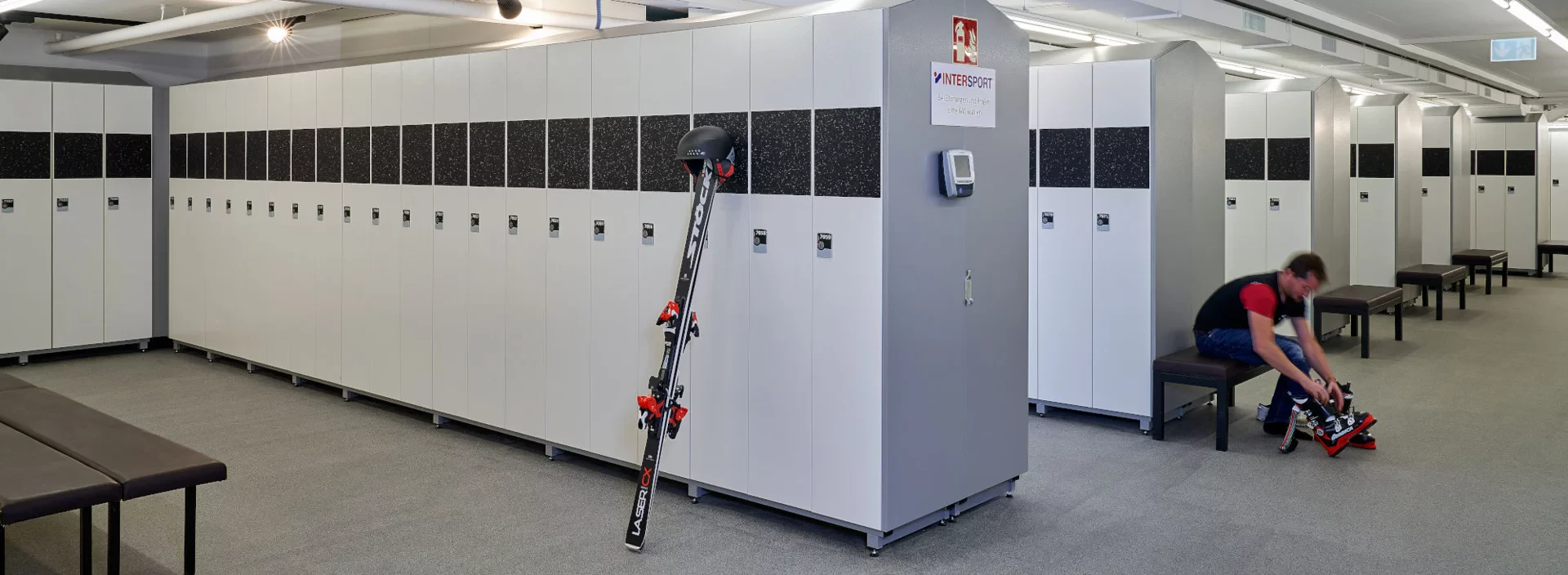A spacious ski locker room with white lockers and benches. A person is sitting, adjusting ski boots. Skis lean against a locker, conveying a calm, organized atmosphere.