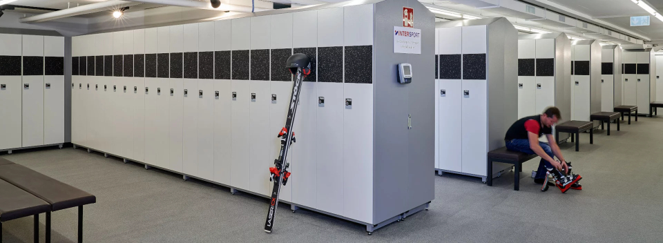 A spacious ski locker room with white lockers and benches. A person is sitting, adjusting ski boots. Skis lean against a locker, conveying a calm, organized atmosphere.