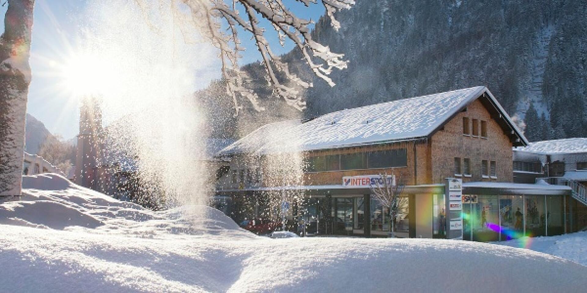 Sonnendurchflutete Winterszene mit schneebedecktem Intersport-Gebäude und Bäumen, Schneeflocken glitzern im Sonnenlicht. Gemütliche, ruhige Atmosphäre, Berge im Hintergrund.