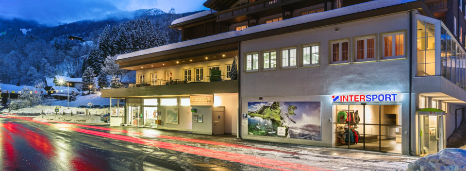 Verschneite Abendstimmung auf der Straße mit einem beleuchteten Intersport-Geschäft. Leuchtende Straßenlaternen spiegeln sich auf dem nassen Asphalt, umgeben von verschneiten Bäumen und Bergen. Gemütliche Atmosphäre.