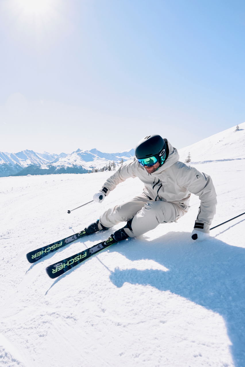 A skier in a pink jacket expertly carves through fresh snow on a mountain slope. Dramatic snowy peaks stand in the misty gray background, conveying action and thrill.