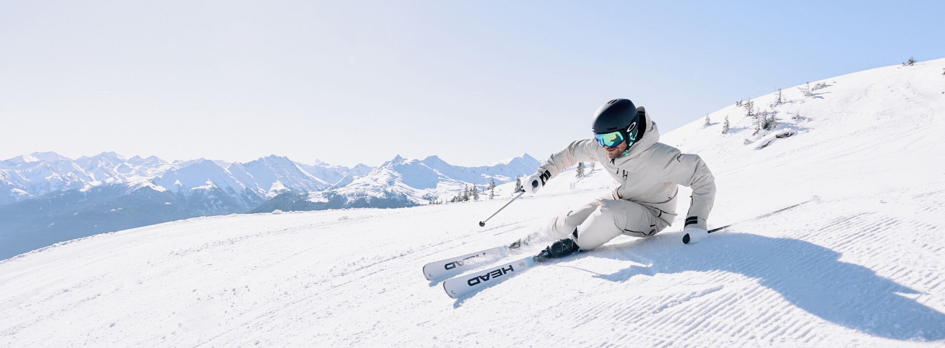 Ein Skifahrer in weißem Anzug und Helm carvt auf einem sonnigen Alpenhang, mit schneebedeckten Bergen in der Ferne.