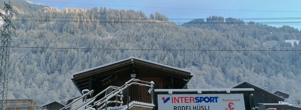 A snowy mountain village scene with a wooden chalet in the foreground. Snow covers the roofs and trees, under a bright, clear sky, conveying a serene winter setting.