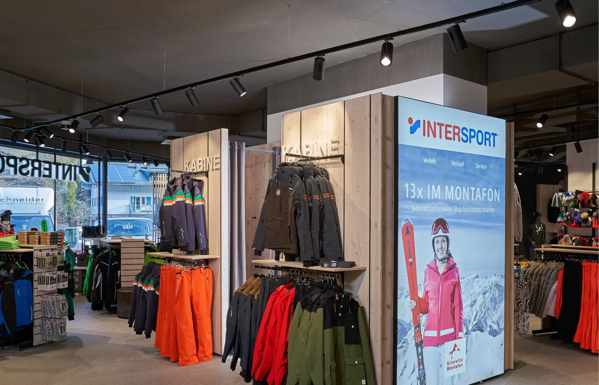 Innenraum eines Einzelhandelsgeschäfts mit Sportbekleidung, in dem farbenfrohe Jacken und Hosen auf Kleiderständern ausgestellt sind. Ein Plakat wirbt für Wintersport und vermittelt eine aktive Outdoor-Atmosphäre.