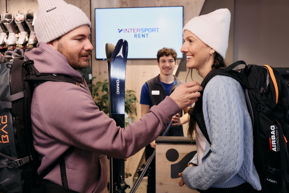Helles Interieur eines Skiverleihgeschäfts mit Holzregalen, Ausstellungsständern und Kassentischen mit digitalen Bildschirmen. Skier und Helme sind gut sichtbar.