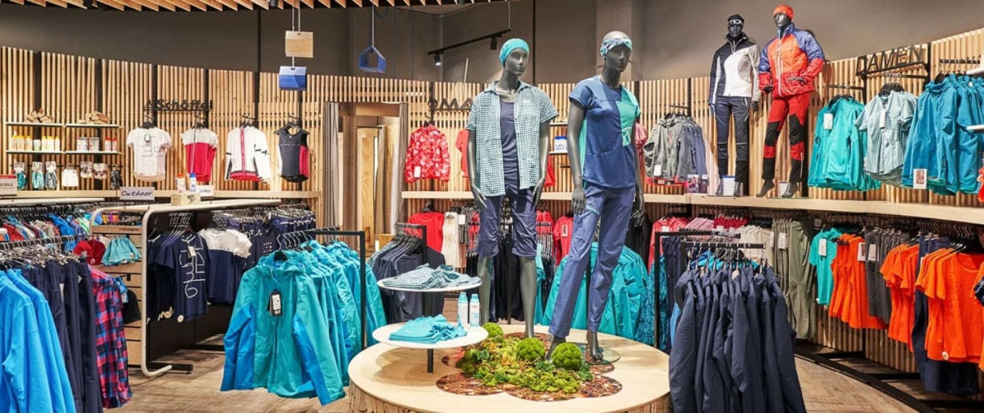 Ein lebhaftes Bekleidungsgeschäft mit Holzakzenten präsentiert farbenfrohe Outdoor-Bekleidung. Mannequins in Sportbekleidung stehen auf einer runden Plattform, umgeben von Kleiderständern mit Hemden und Jacken.