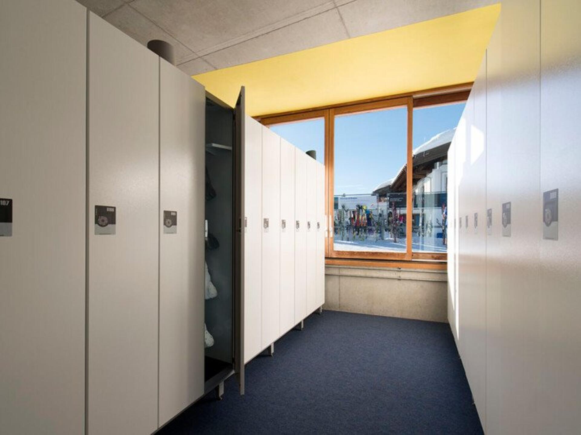 Heller Umkleideraum mit weißen Spinden, eine Tür steht offen und gibt den Blick auf die darin befindliche Kleidung frei. Ein großes Fenster bietet einen Blick nach draußen auf Menschen und ein modernes Gebäude.