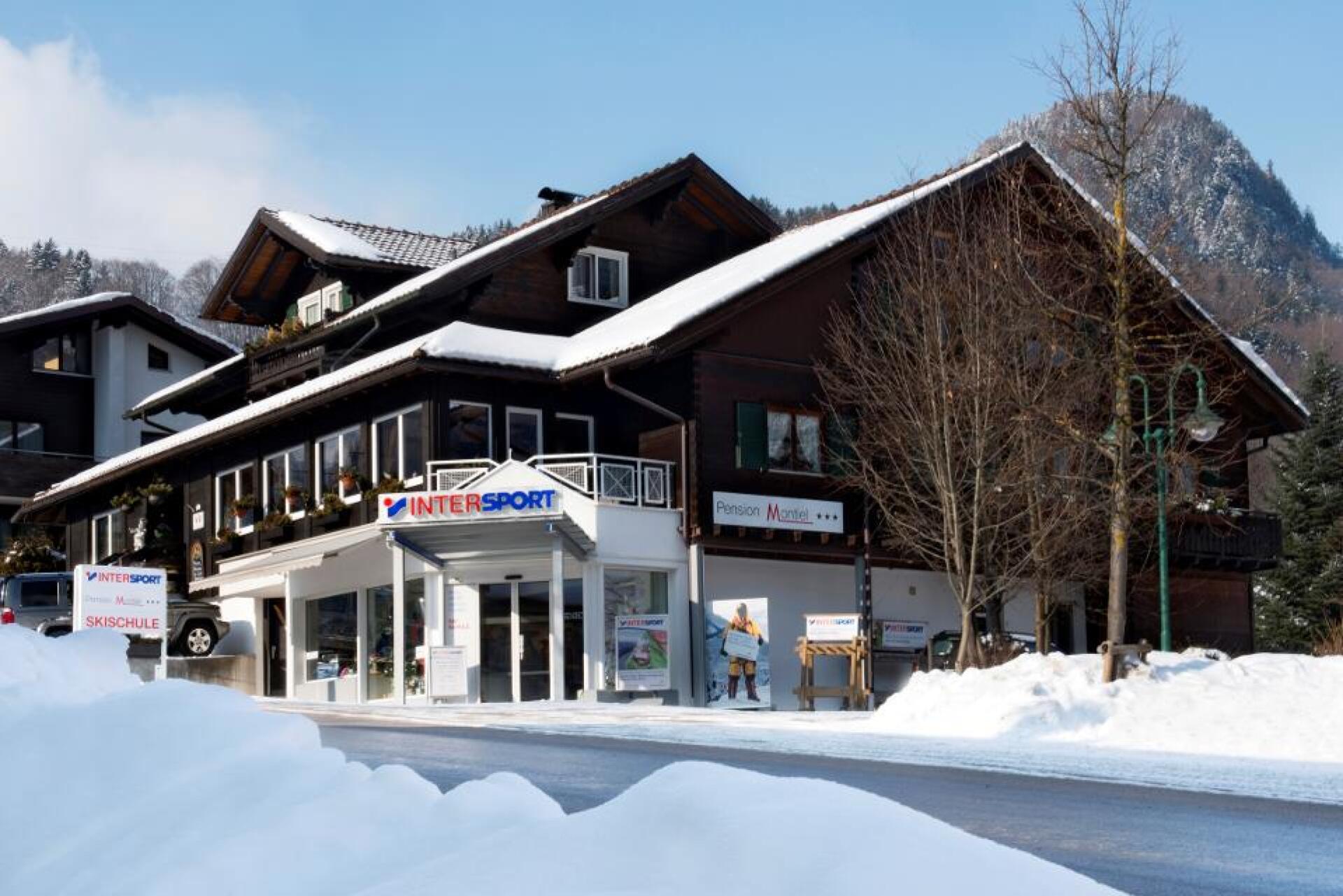 A snow-covered chalet-style building houses a ski shop and pharmacy, set against a mountainous backdrop. The scene conveys a tranquil winter atmosphere.