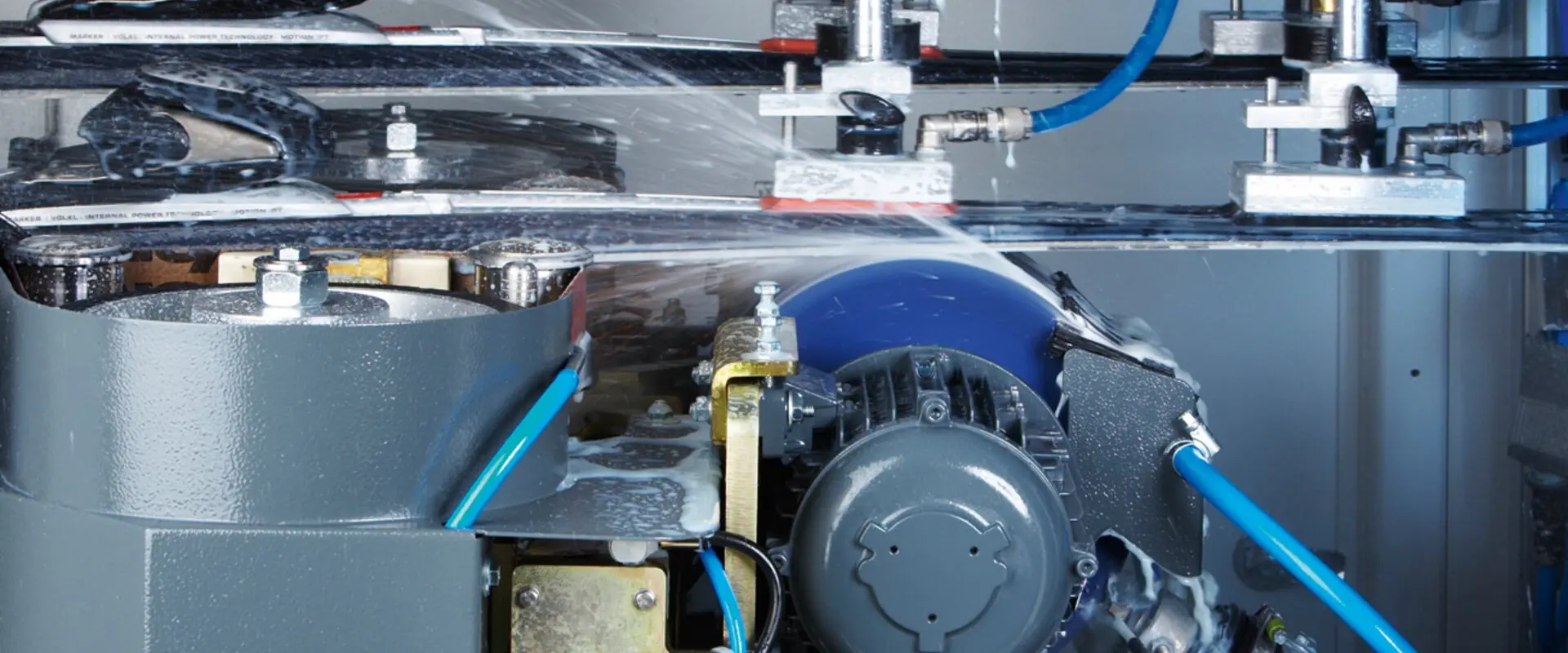 Close-up of an industrial machine in operation, featuring metallic parts, blue cables, and water spraying, creating a dynamic and technical atmosphere.
