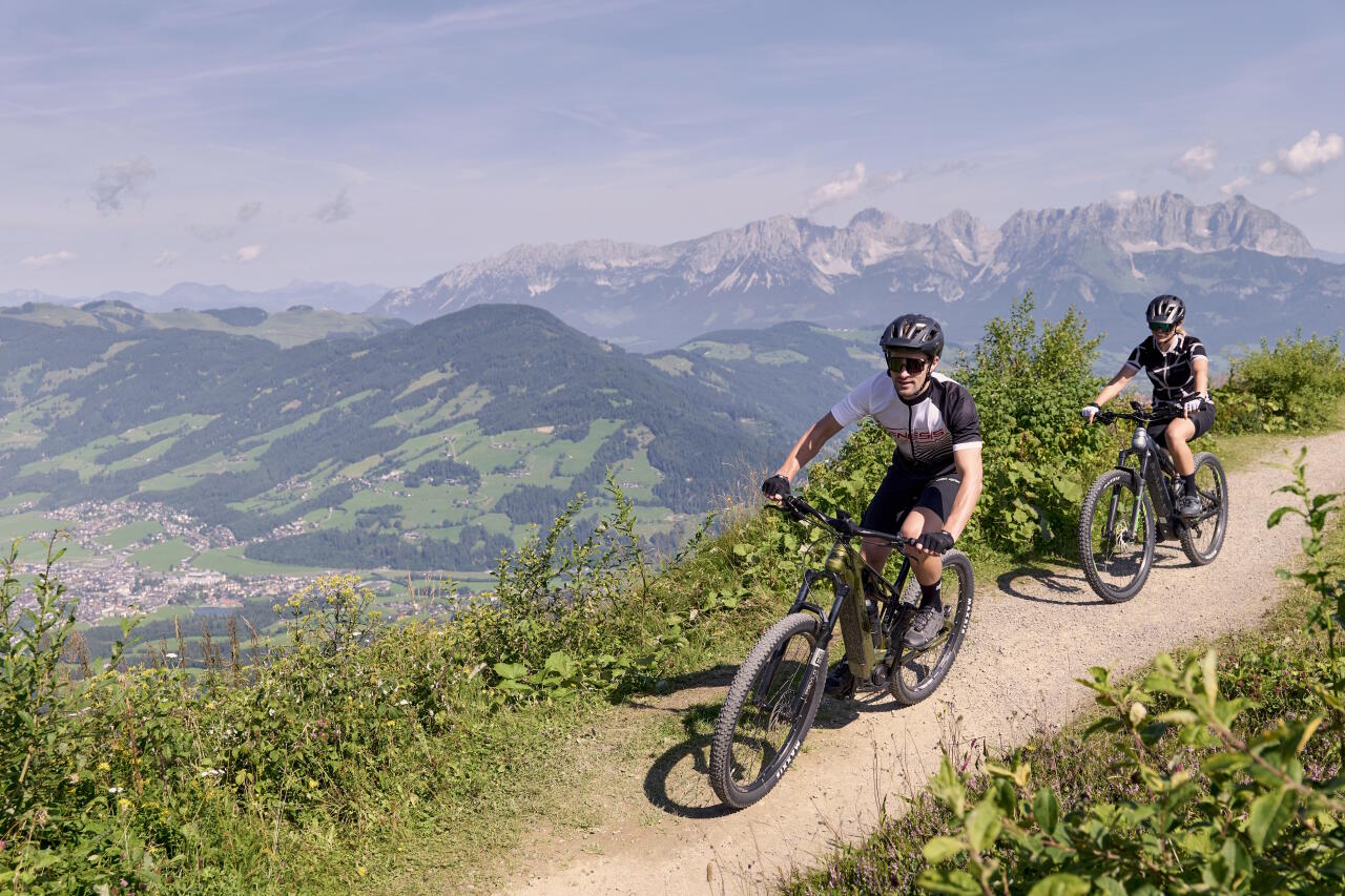 Two cyclists on mountain bikes ride along a sunny mountain trail with a view over the green valley and the Alps in the background.