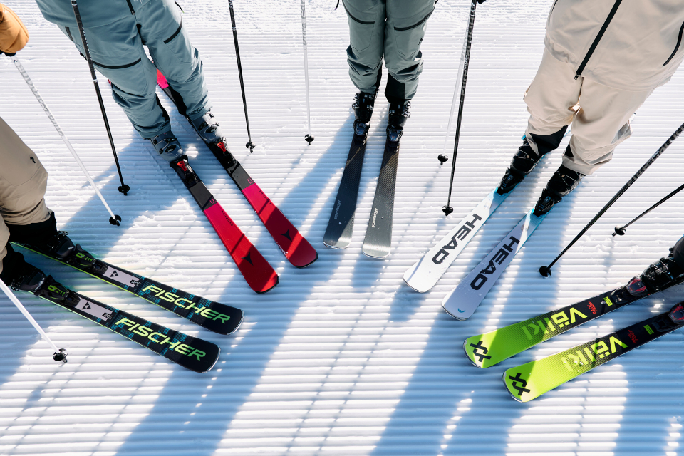 A person in white pants holds an orange ski horizontally across their waist. The wooden background and shadows convey a relaxed, stylish atmosphere.