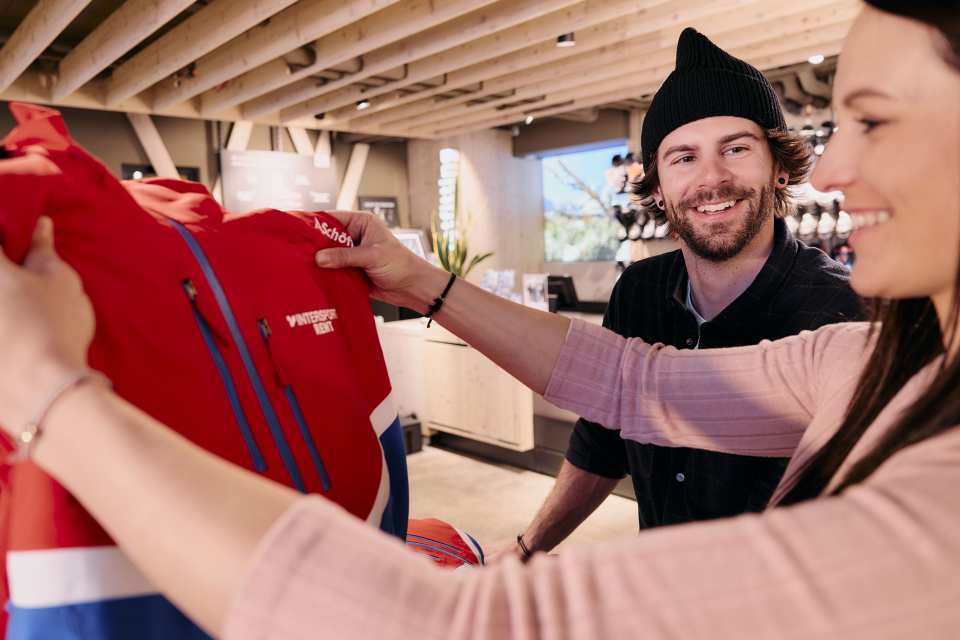 Ein gut beleuchtetes Bekleidungsgeschäft mit Regalen voller bunter Jacken, Hemden und Outdoor-Ausrüstung auf beiden Seiten. Der Raum wirkt aufgeräumt und einladend.