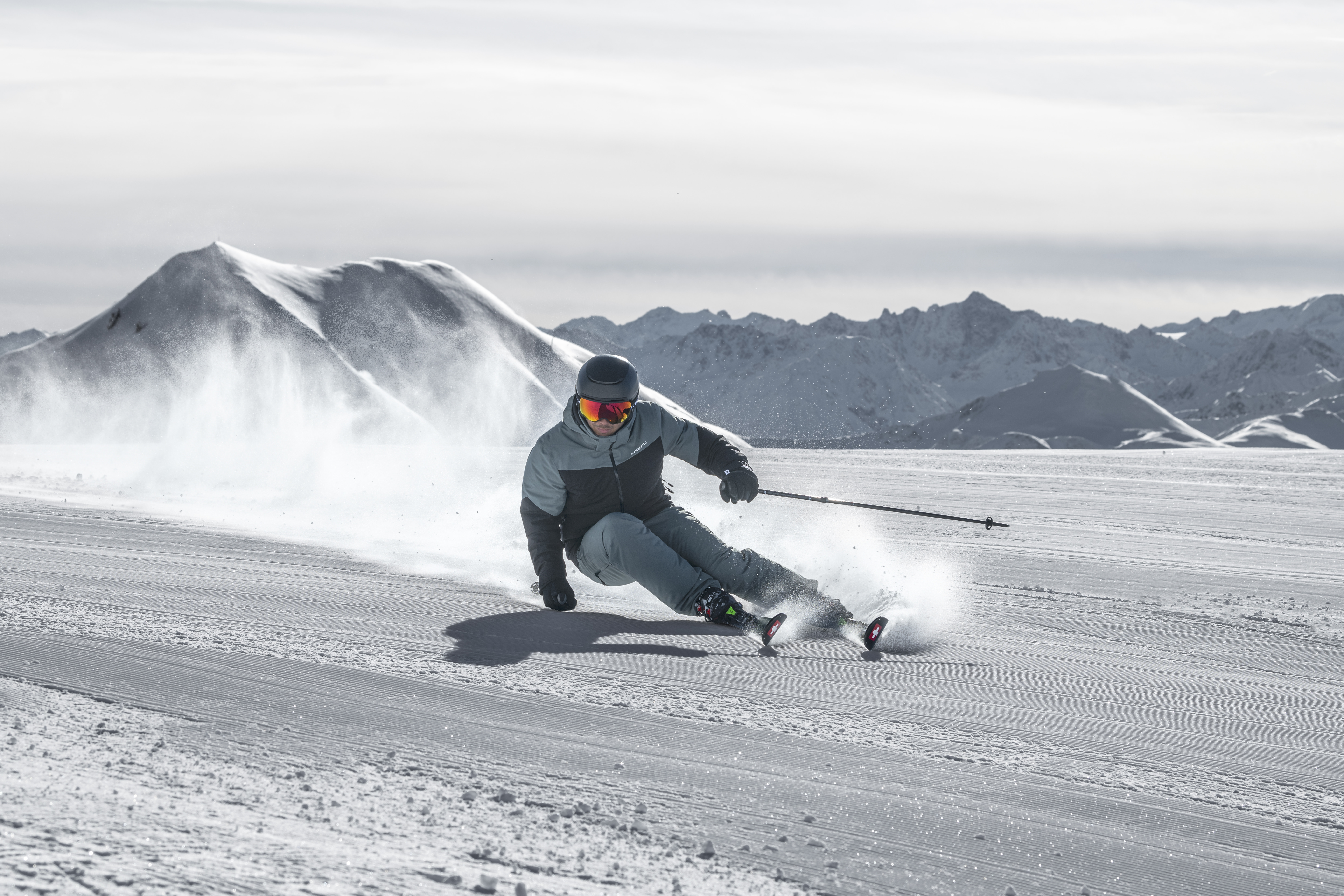 Ein Skifahrer in einer rosa Jacke carvt gekonnt durch den Neuschnee auf einem Berghang. Dramatische schneebedeckte Gipfel ragen vor dem nebligen grauen Hintergrund empor und vermitteln Action und Nervenkitzel.