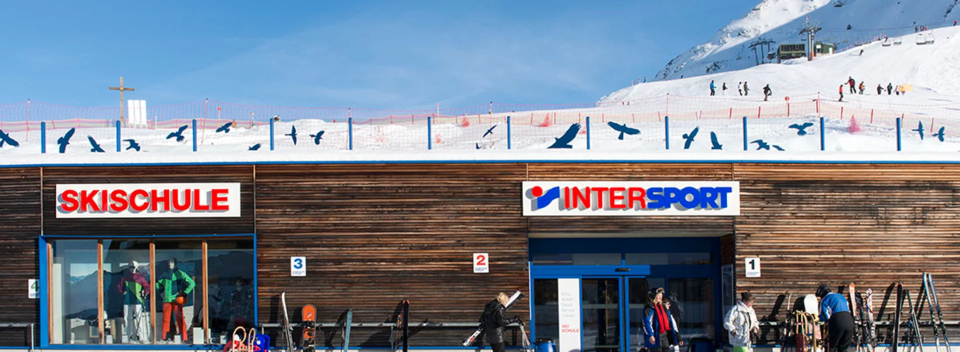 Wooden building with “Skischule” and “Intersport” signs. Ski equipment outside. Snowy slopes and skiers visible in the background, bright sky.