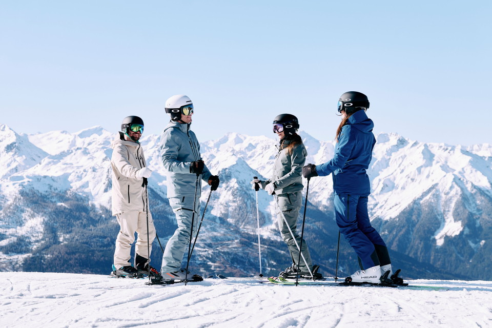 Drei Skifahrer in farbenfroher Ausrüstung heben freudig ihre Skistöcke vor einem strahlend blauen Himmel. Im Hintergrund erstrecken sich schneebedeckte Berge, die Spannung und Abenteuer versprechen.