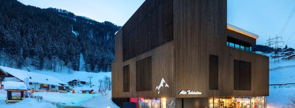 Modernes Holzgebäude in der Abenddämmerung in einem verschneiten Skidorf, umgeben von schneebedeckten Bäumen und Bergen, das eine ruhige Winteratmosphäre vermittelt.