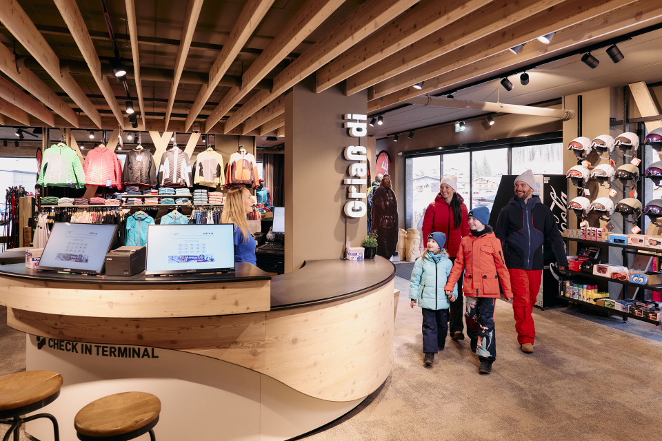 A spacious ski rental shop features wooden shelves with colorful skis and poles. Counters with monitors are aligned neatly; ski boots rest on the floor.