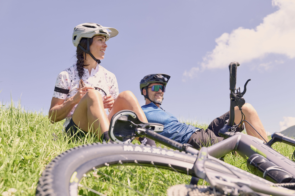 Drei Wanderer ruhen sich auf felsigem Gelände aus, gekleidet in farbenfrohe Kleidung, vor dem Hintergrund schneebedeckter Berge unter einem bewölkten Himmel, und vermitteln dabei ein Gefühl von Abenteuer und Kameradschaft.
