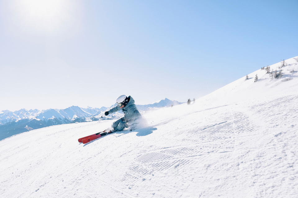 Ein Skifahrer rast vor dem Hintergrund sonnenbeschienener Berge einen verschneiten Hang hinunter und vermitteln dabei Energie und Begeisterung.